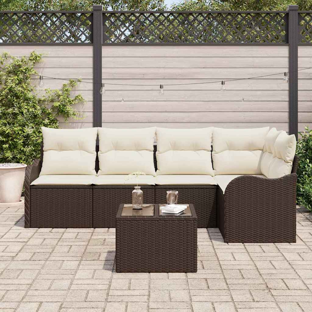 Garten-Terrassenset. Braunes Korbmöbelsofa mit cremefarbenen Kissen und einem Couchtisch mit Glasplatte. Holzzaun im Hintergrund.