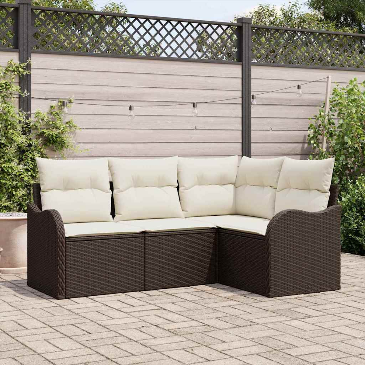 Ein Ecksofa im Freien mit cremefarbenen Kissen steht auf einer Steinterrasse. Über dem Sofa hängt ein Zaun mit Lichtern, im Hintergrund Grün.