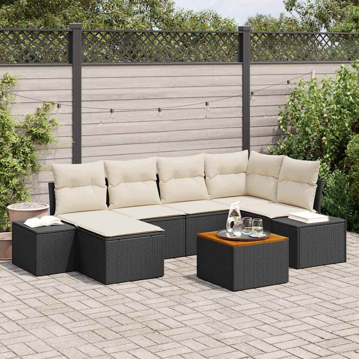 Garten-Sitzgruppe auf einer Terrasse. Sie umfasst ein Ecksofa mit cremefarbenen Kissen, einen Couchtisch und zwei Beistelltische. Im Hintergrund ein Zaun.