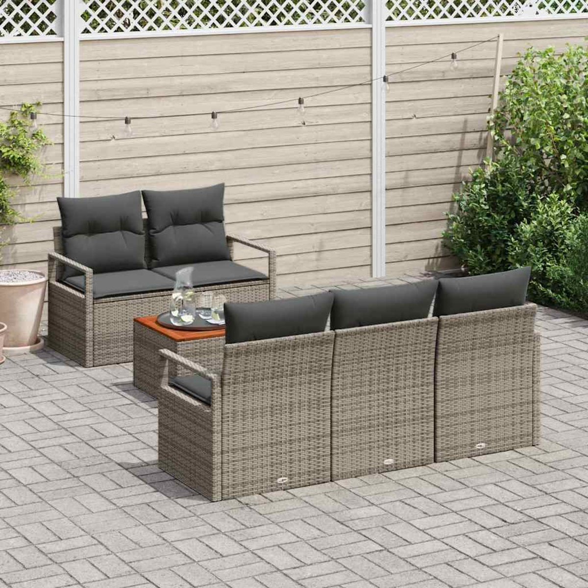 Garten-Terrassenset mit grauen Kissen auf grauem Korbbasis. Kleiner Tisch mit Gläsern. Hintergrund Zaun und Grün.