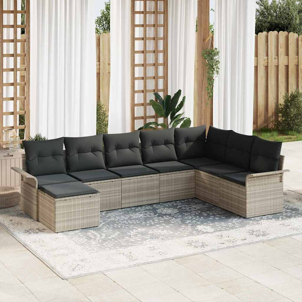 Ein Ecksofa im Freien mit dunkelgrauen Kissen auf einem gemusterten Teppich, in der Nähe weißer Vorhänge und Holzgitter, mit Pflanzen und Zaun.