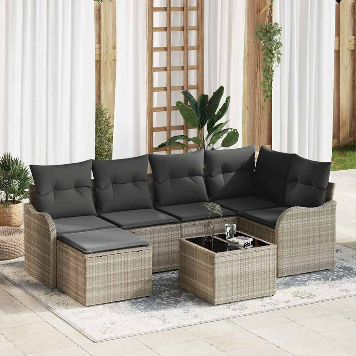 Rattan-Sofa im Freien mit grauen Kissen und einem Couchtisch mit Glasplatte auf einem gemusterten Teppich, mit Vorhängen und Pflanzen im Hintergrund.