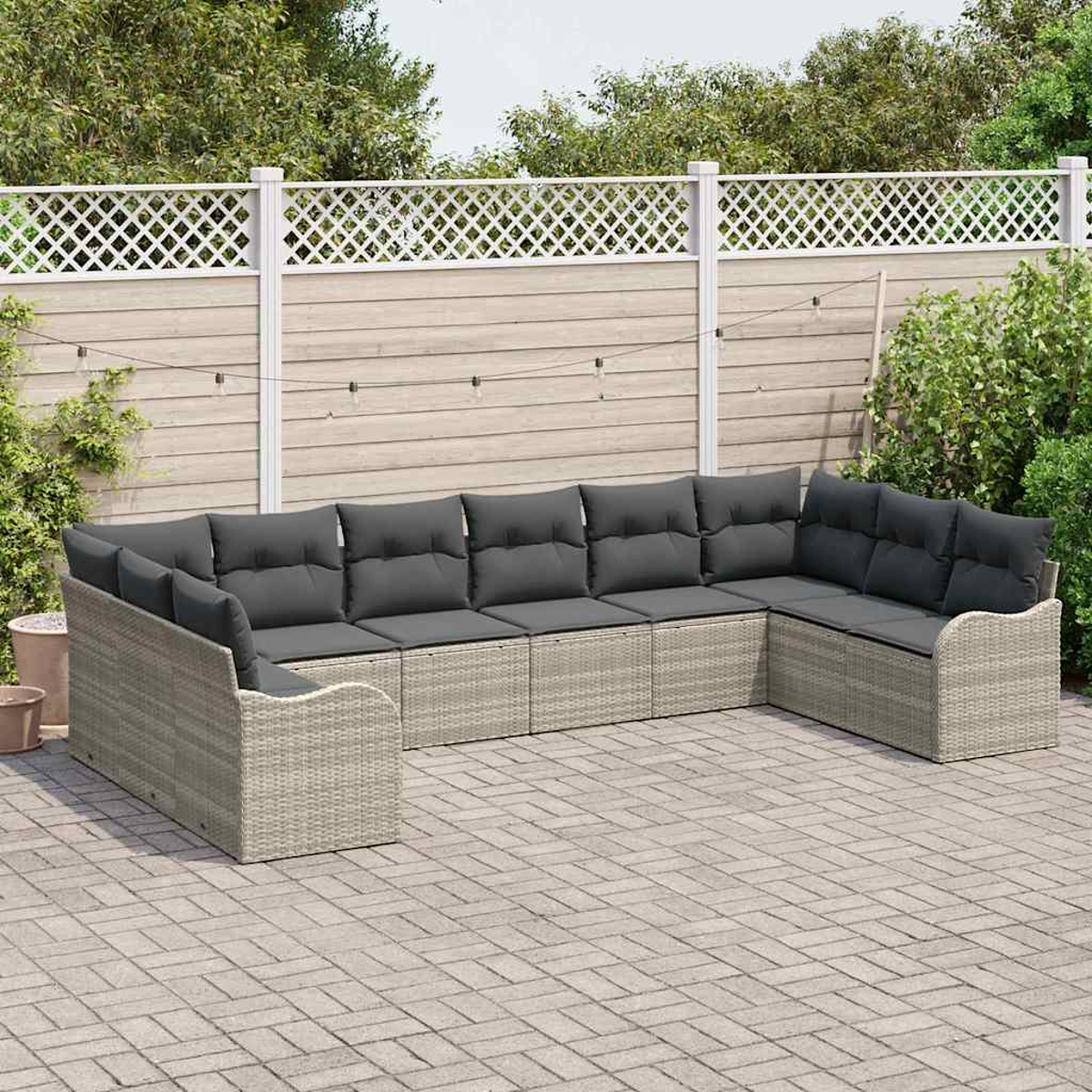Ein graues Rattan-Ecksofa mit dunkelgrauen Kissen steht auf einer Terrasse. Ein weißer Zaun und Grünzeug sind im Hintergrund.