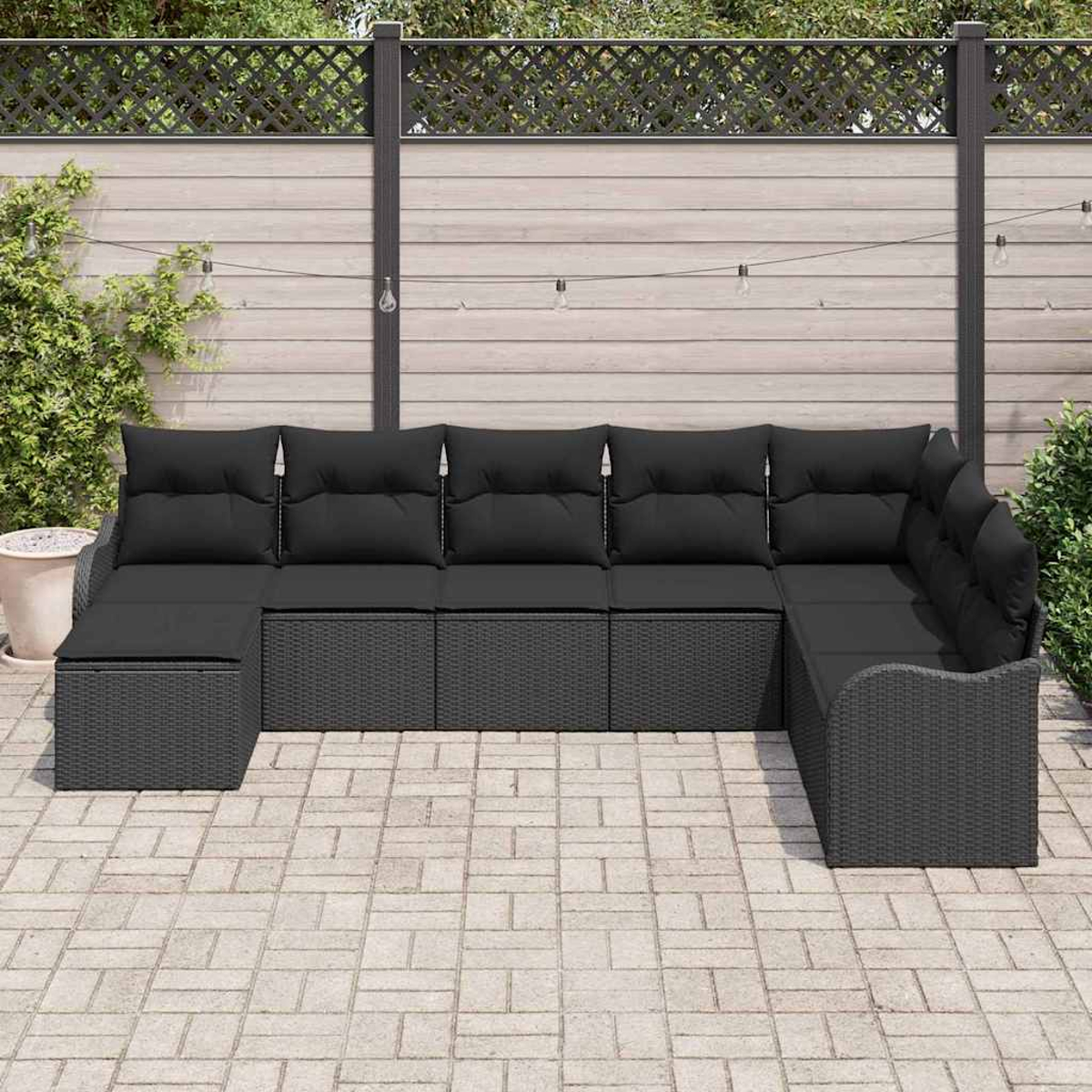 Ein schwarzes Outdoor-Ecksofa aus Korbgeflecht mit Kissen steht auf einer Terrasse. Im Hintergrund ein Holzzaun mit Rankgitter.