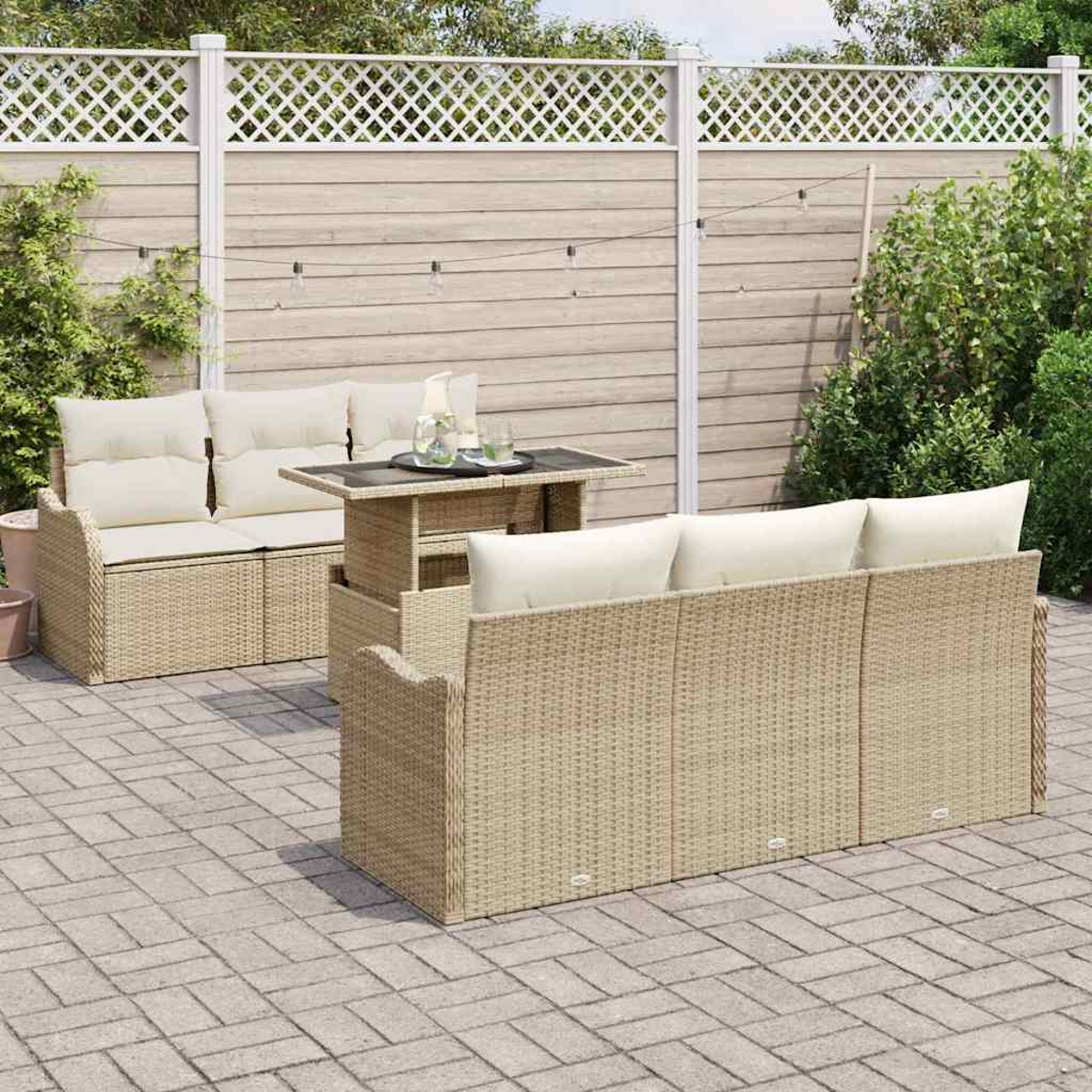 Gartenmöbel-Set aus Rattan mit beigen Kissen und einem Tisch auf einer Ziegelstein-Terrasse. Ein Holzzaun und Grünzeug im Hintergrund.