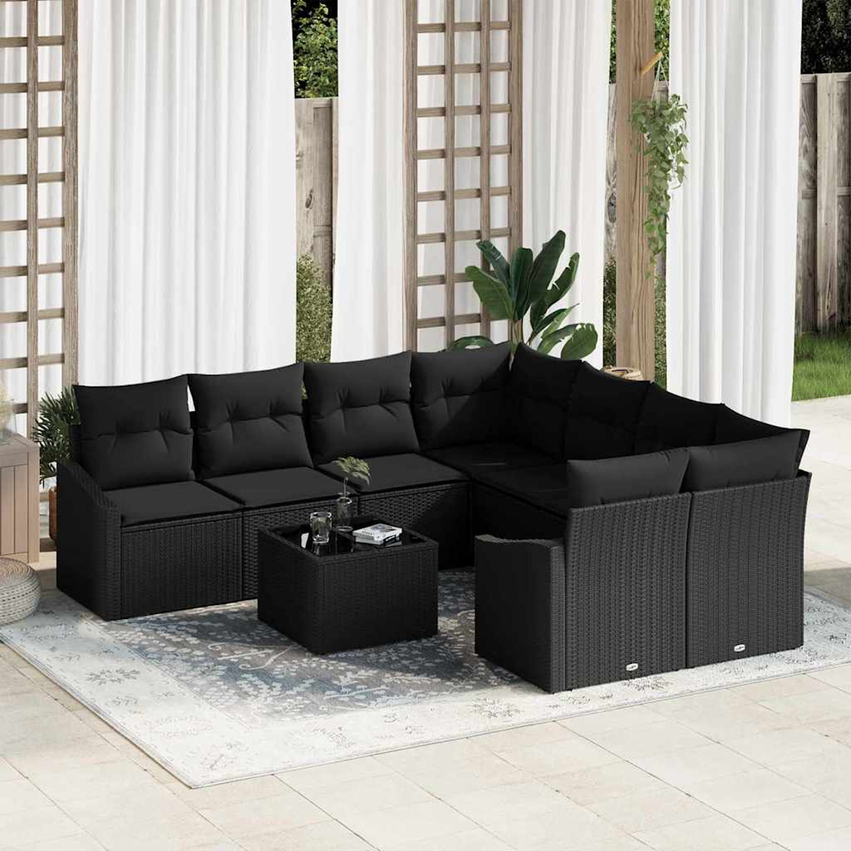 Sofá seccional de mimbre negro para exteriores con cojines, una mesa de centro y una alfombra estampada. Cortinas blancas transparentes al fondo.