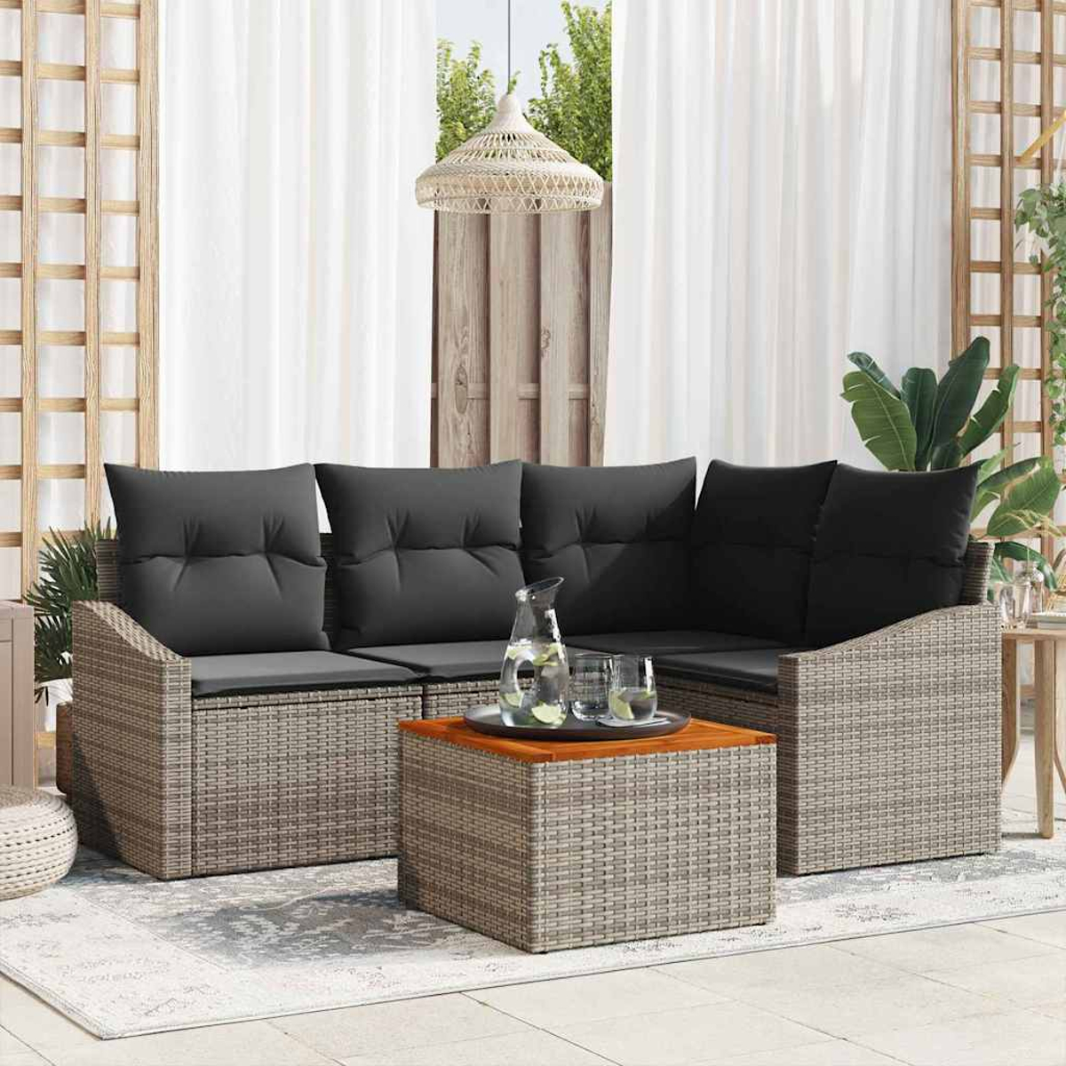Sofá de rat án exterior con cojines grises, mesa de centro con vasos y jarra. Cortinas blancas y plantas al fondo.