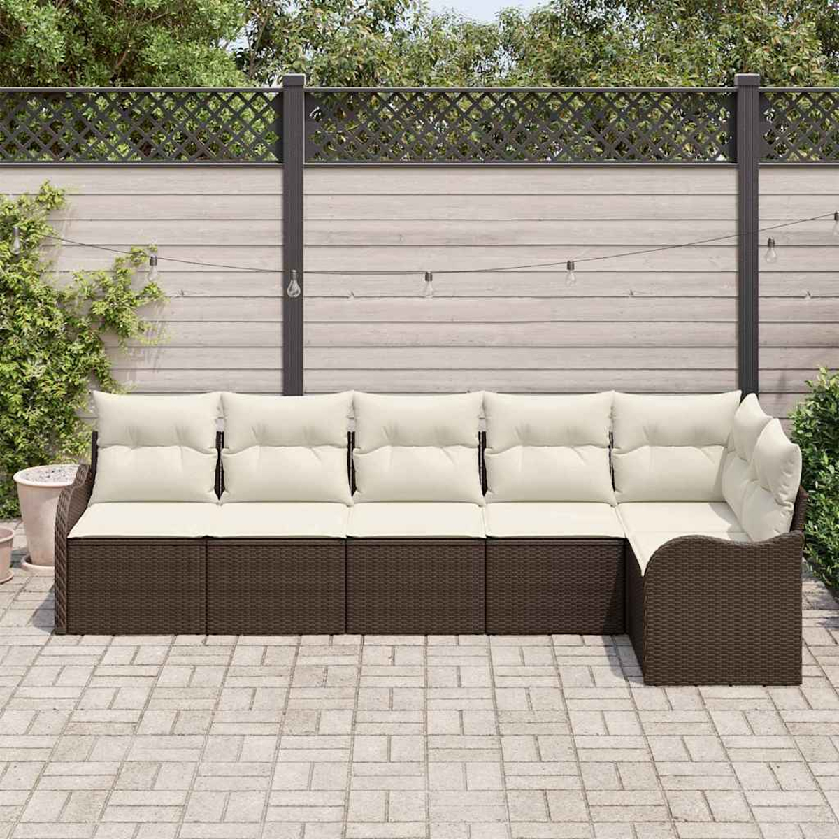 Ein Ecksofa für den Außenbereich mit cremefarbenen Kissen und braunem Korbgewebe. Es steht auf einer Terrasse vor einem Holzzaun und Grün.