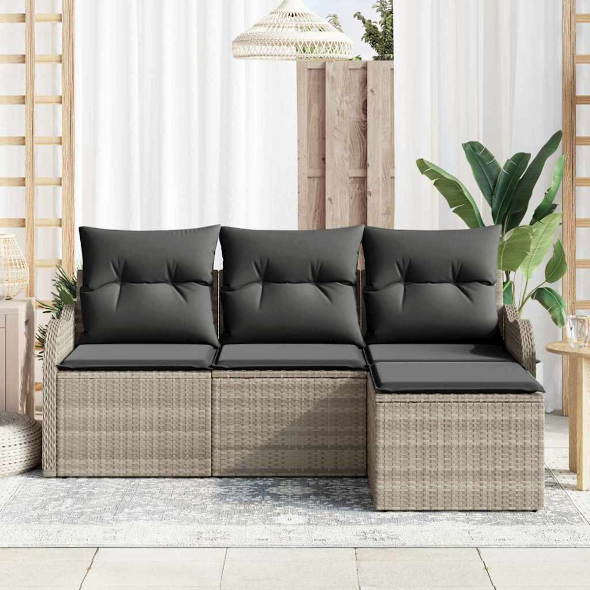 Ein Rattansofa mit dunkelgrauen Kissen steht auf einer Terrasse. Im Hintergrund befinden sich ein weißer Vorhang und eine Pflanze.