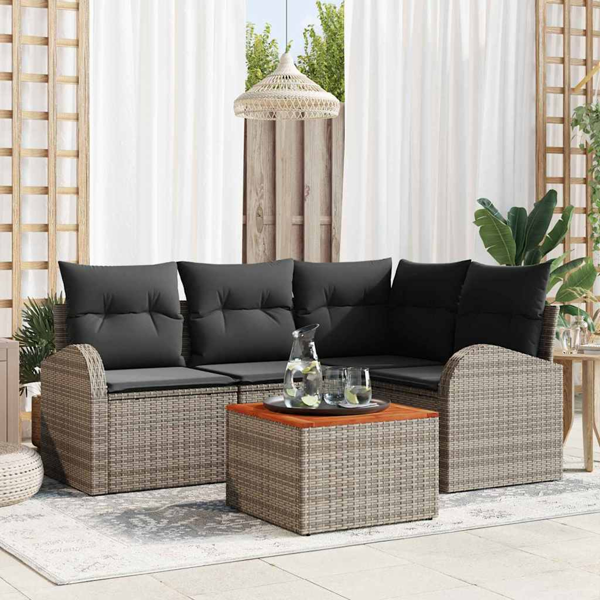 Outdoor-Sitzbereich mit grauen Kissen, Rattanmöbeln, Holztisch und weißen Vorhängen. Eine Kanne und Gläser stehen auf dem Tisch.