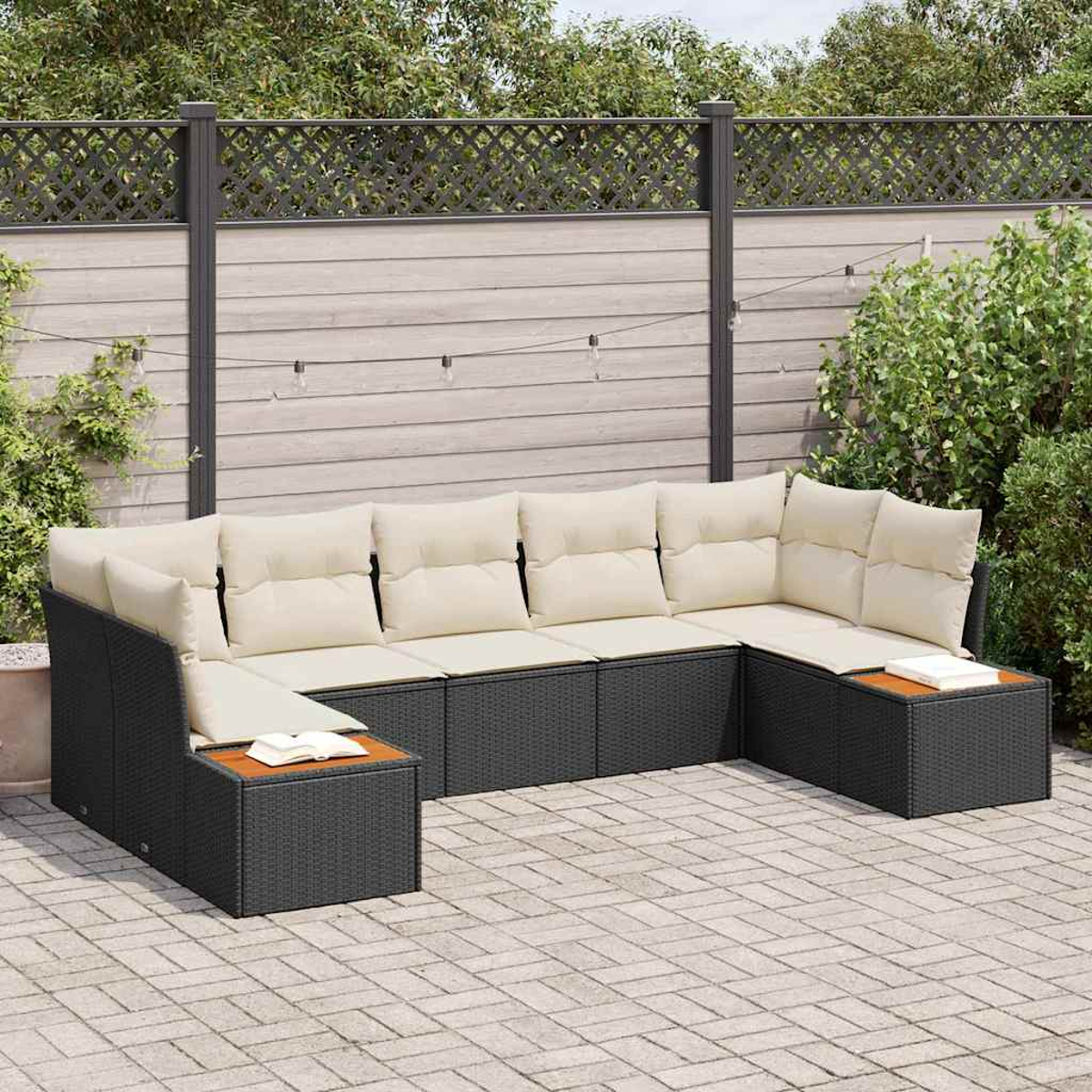 Sofá seccional en forma de U para exteriores con cojines crema y estructura de mimbre gris oscuro. Mesas laterales de madera con libros. Fondo: valla de madera y plantas.