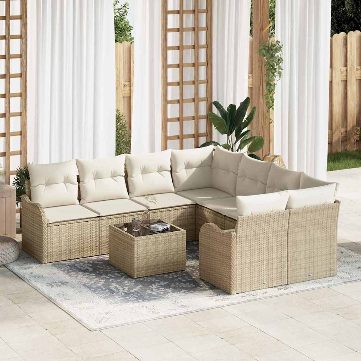 Outdoor-Sofa aus Korbgeflecht mit cremefarbenen Kissen und einem passenden Couchtisch auf einem gemusterten Teppich, vor weißen Vorhängen.