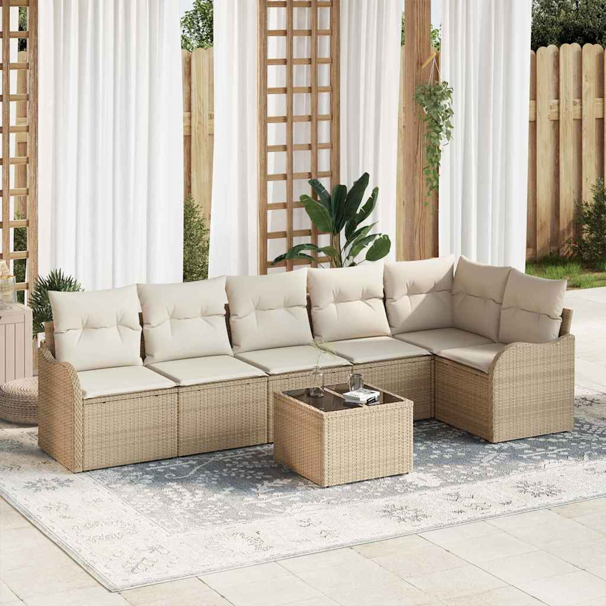Ein Outdoor-Sofa aus Korbgeflecht mit beigen Kissen steht auf einem gemusterten Teppich. Ein kleiner Tisch und weiße Vorhänge sind im Hintergrund.