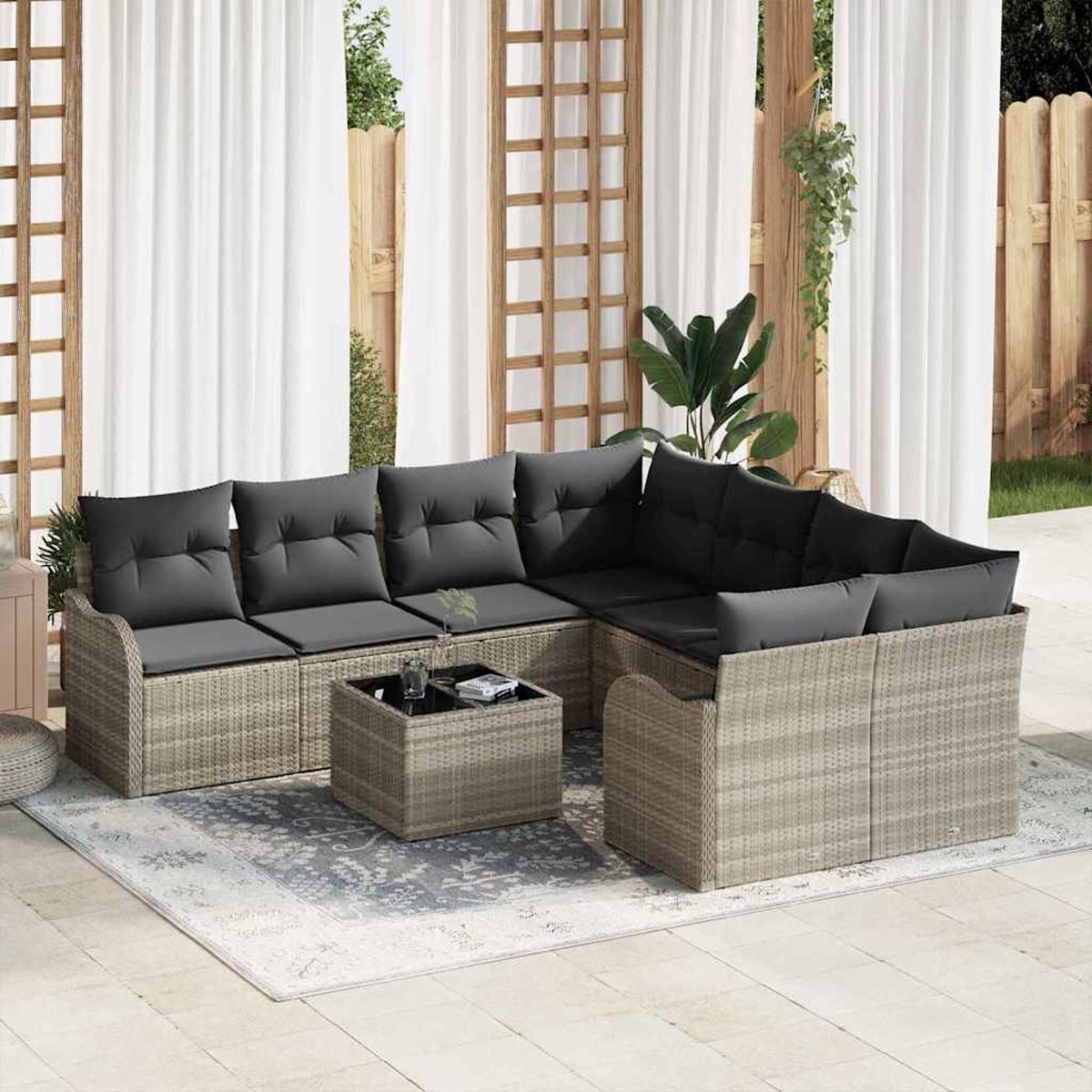 Outdoor-Sitzgruppe mit grauen Kissen und gewebtem Rahmen. Ein kleiner Tisch steht auf einem gemusterten Teppich. Weiße Vorhänge und ein Holzzaun sind im Hintergrund.