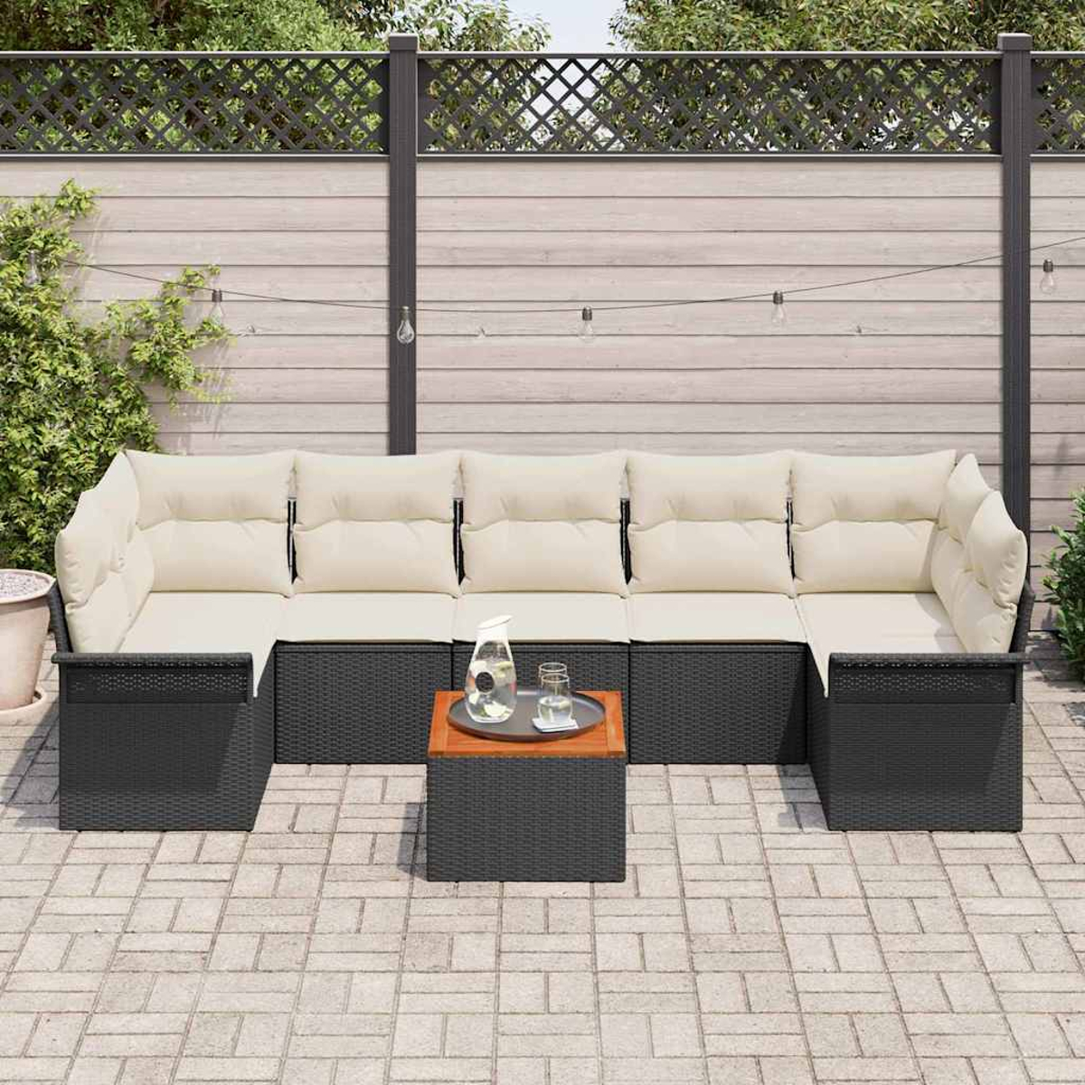 Sofá seccional exterior con cojines beige y estructura de ratán negro. Mesa de café de madera. Valla de madera al fondo, luces.