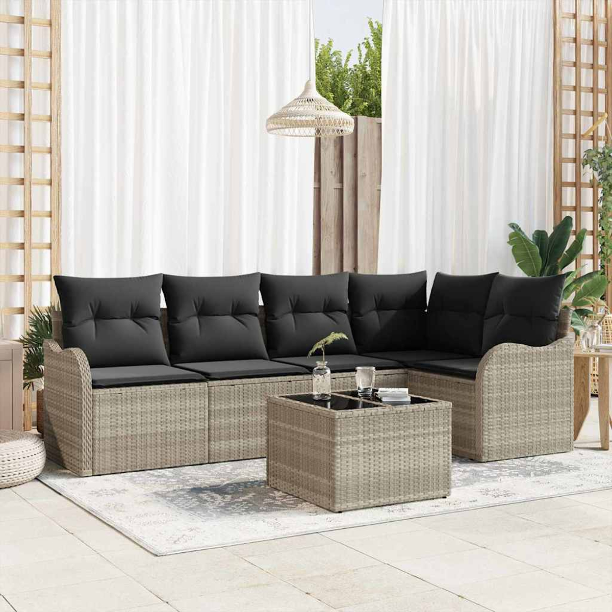 Rattansofa mit schwarzen Kissen und einem quadratischen Tisch auf einem Teppich. Im Hintergrund weiße Vorhänge und grüne Pflanzen.