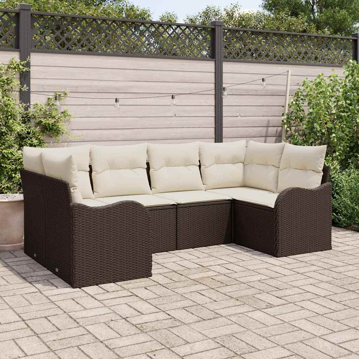 Ein braunes Korbsessel-Sofa mit cremefarbenen Kissen steht auf einer Terrasse. Im Hintergrund ein Holzzaun mit Lichterketten und Grün.