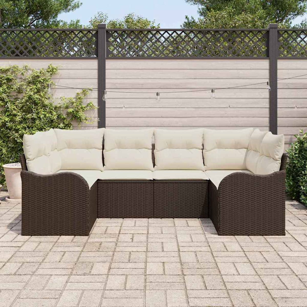 Ein braunes Korbsessel-Sofa mit cremefarbenen Kissen steht auf einer Backstein-Terrasse. Ein Holzzaun und Grünzeug sind im Hintergrund.