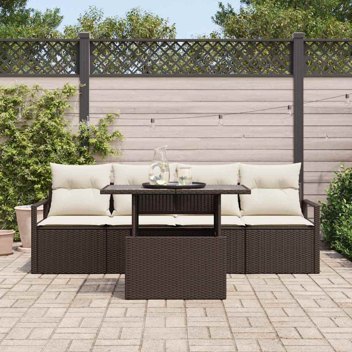 Gartenmöbel mit dunkelbraunem, gewebtem Rahmen, cremefarbenen Kissen und einem passenden Tisch. Der Hintergrund umfasst einen Holzzaun und Grün.
