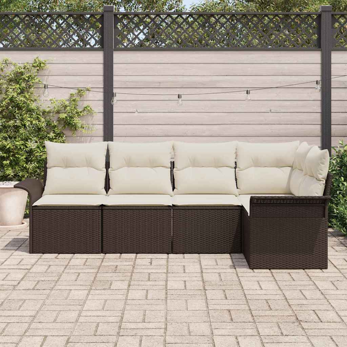Ein braunes Outdoor-Ecksofa aus Korbgeflecht mit cremefarbenen Kissen steht auf einer Backstein-Terrasse. Dahinter ist ein Holzzaun mit Weinreben.