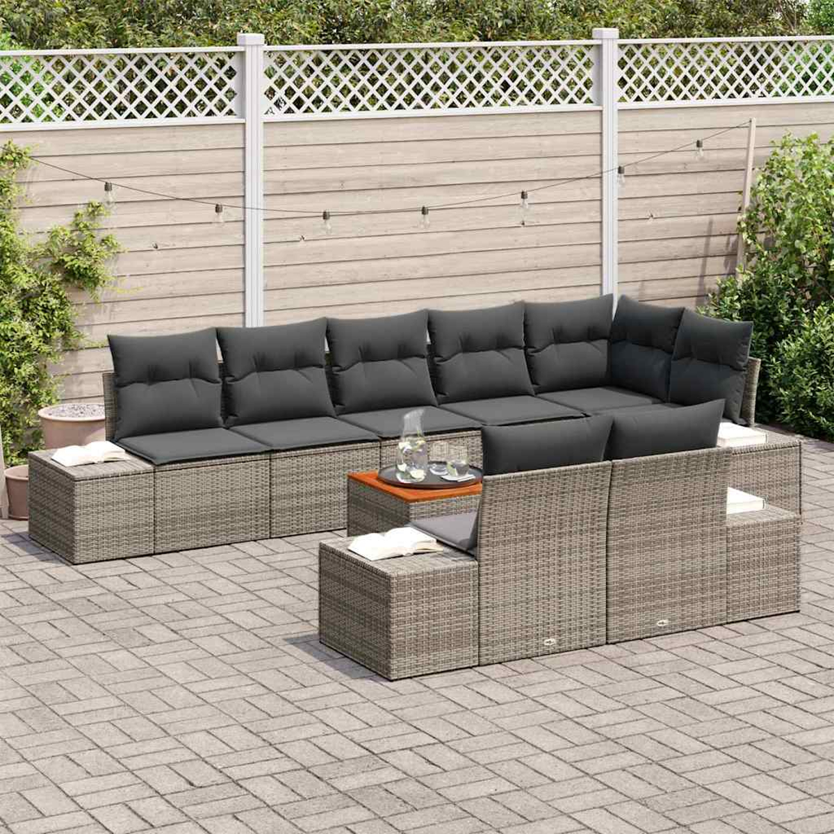 Garten-Terrassenset mit grauen Kissen. Holzzaun im Hintergrund. Tisch mit Gläsern. Ziegelpflaster. Grüne Pflanzen an den Seiten.