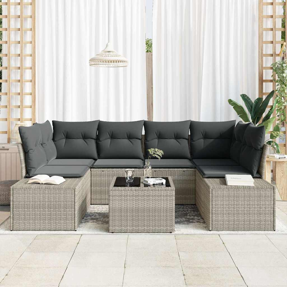 Sofá seccional exterior con cojines y almohadas grises, pequeñas mesas cuadradas y cortinas blancas al fondo. Hay un libro en el sofá.