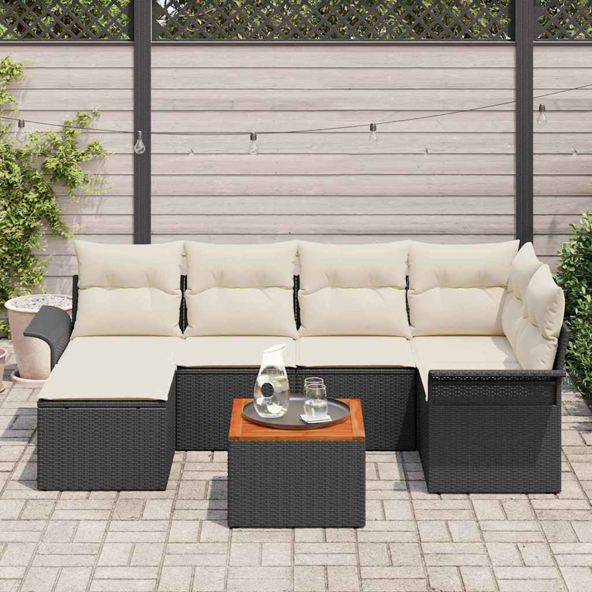 Outdoor-Ecksofa mit cremefarbenen Kissen, schwarzem Korbgewebe. Kleiner Holztisch mit Glaskrug und Gläsern. Ziegelterrasse im Vordergrund.