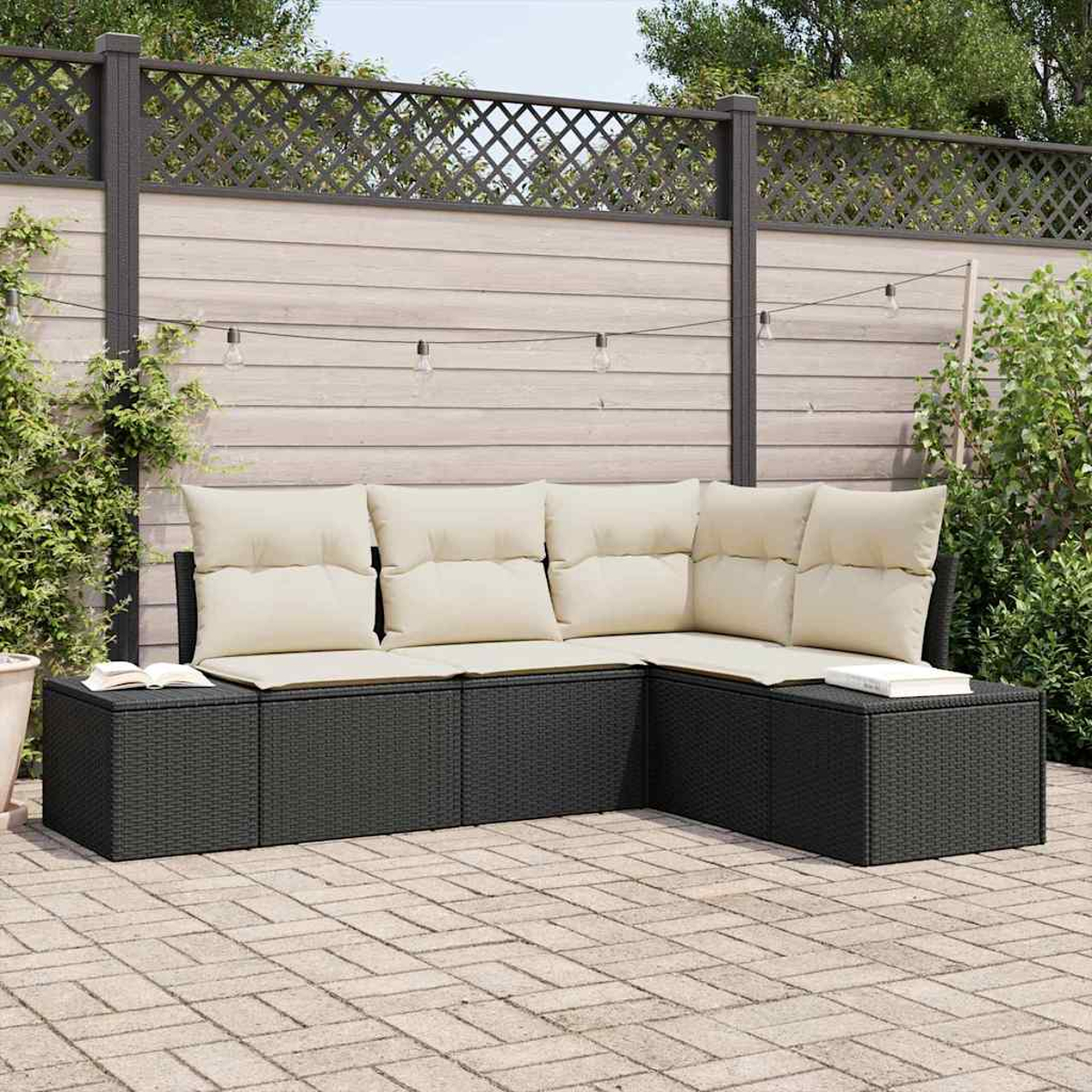 Outdoor-Ecksofa mit cremefarbenen Kissen. Schwarze Korbmöbel auf einer gepflasterten Terrasse. Holzzaun mit Lichterketten im Hintergrund.