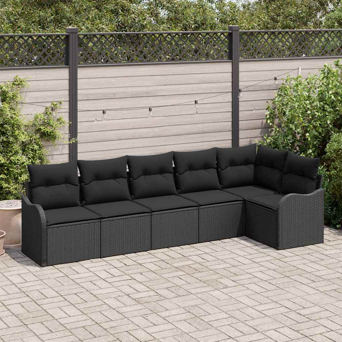 Ein schwarzes Korbsessel-Ecksofa mit schwarzen Kissen steht auf einer Terrasse. Ein Holzzaun und Grünzeug sind im Hintergrund.