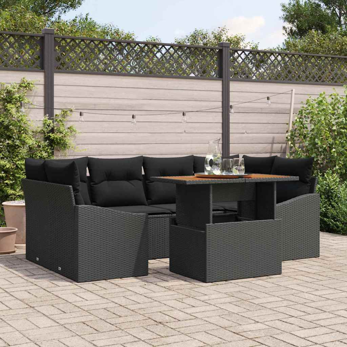 Garten-Terrassenset mit schwarzen Kissen und einem Holztisch. Auf dem Tisch stehen ein klarer Krug und Gläser. Im Hintergrund ist ein Zaun.