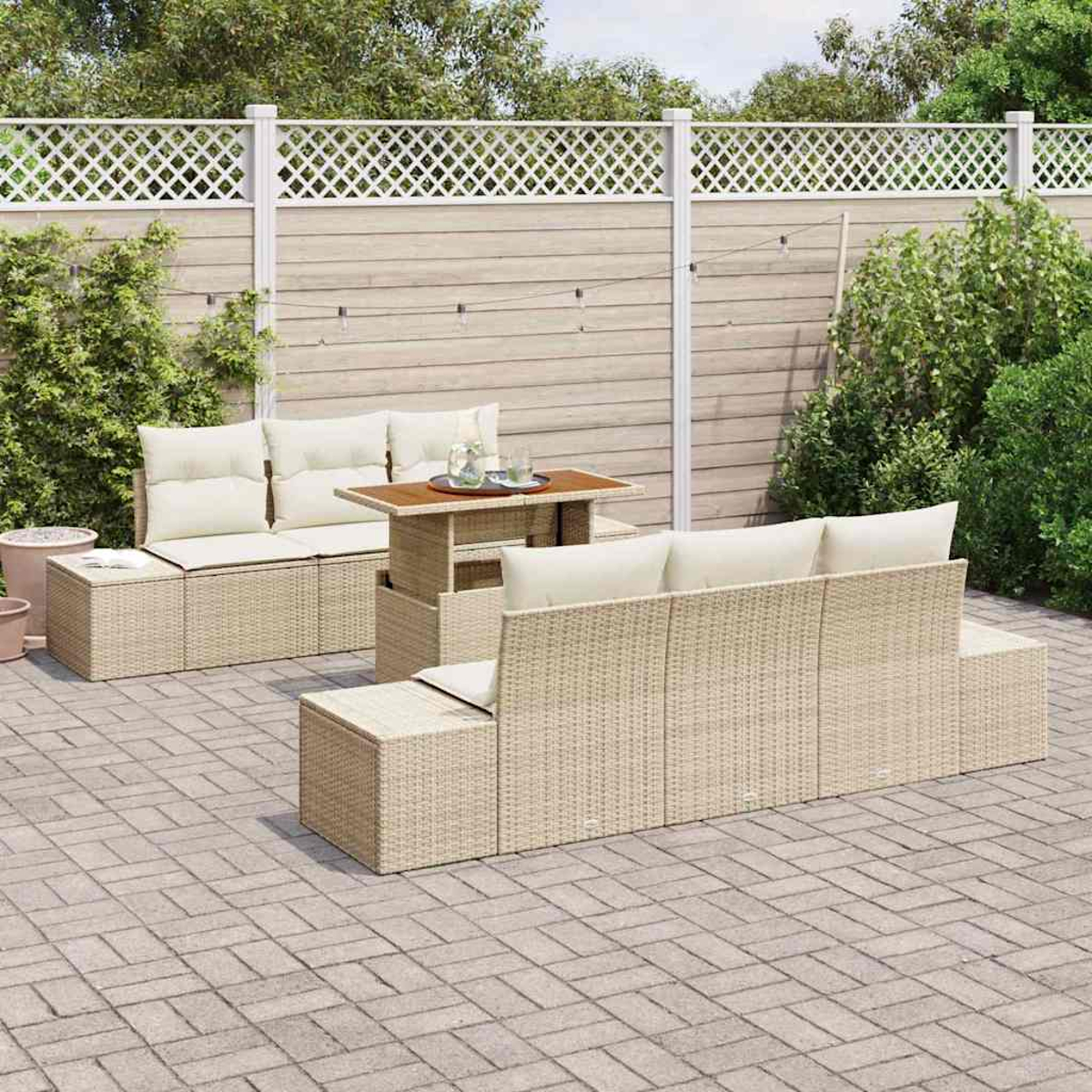 Outdoor-Korbmöbel mit beigen Kissen auf einer Backstein-Terrasse. Ein kleiner Tisch trägt Gläser. Ein weißer Zaun und grüne Pflanzen im Hintergrund.
