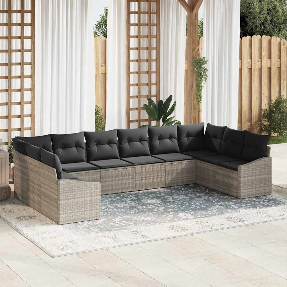 Ein Sitzbereich im Freien mit einem grauen Ecksofa, schwarzen Kissen und einem gemusterten Teppich. Weiße Vorhänge und ein Holzzaun sind im Hintergrund.