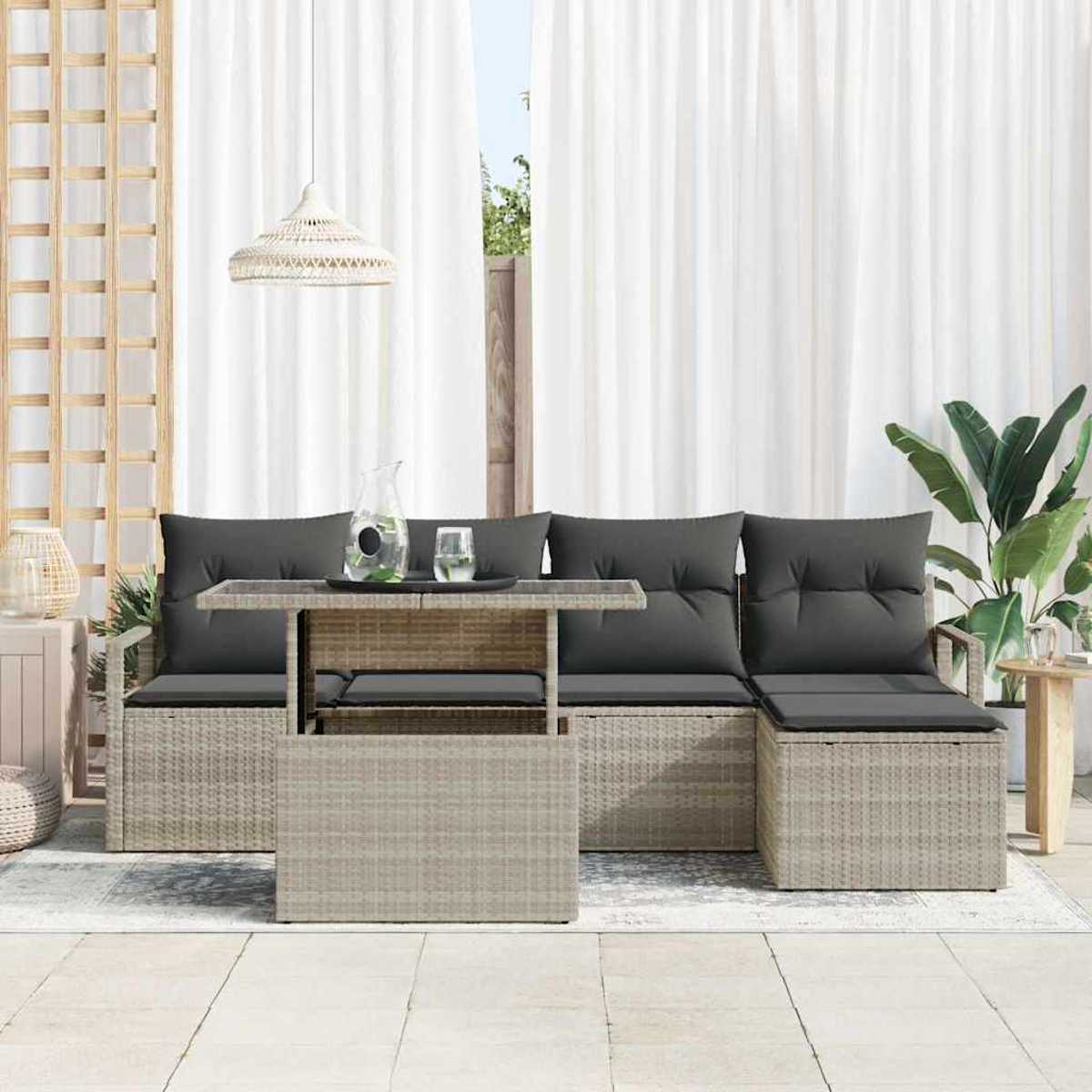 Outdoor-Sitzbereich mit grauen Kissen, einem Tisch mit Glasplatte, einer Wasserkaraffe und einer Pflanze. Weiße Vorhänge im Hintergrund.