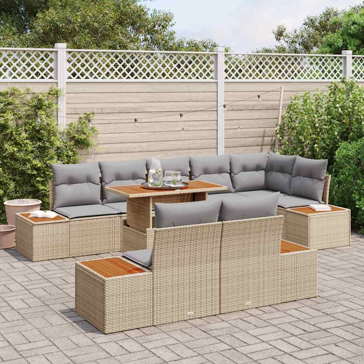 Gartenmöbel-Set aus Rattan mit grauen Kissen und Holztischplatten. Auf einer Backstein-Terrasse, mit weißem Zaun und Grün im Hintergrund.