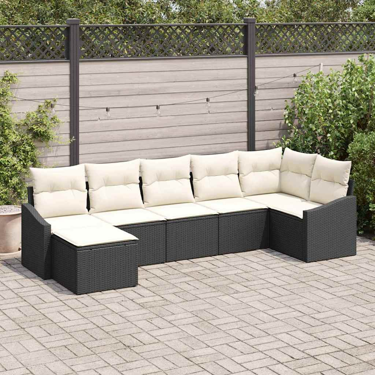 Ein graues Outdoor-Ecksofa aus Korbgeflecht mit weißen Kissen auf einer Ziegelterrasse. Im Hintergrund ein Zaun und Grün.