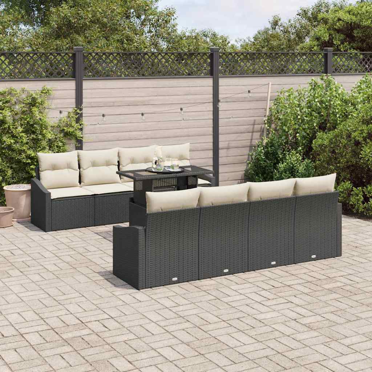 Escena al aire libre con sofás de mimbre negros, cojines beige, una mesa y un patio de ladrillos. Una valla de madera y vegetación al fondo.