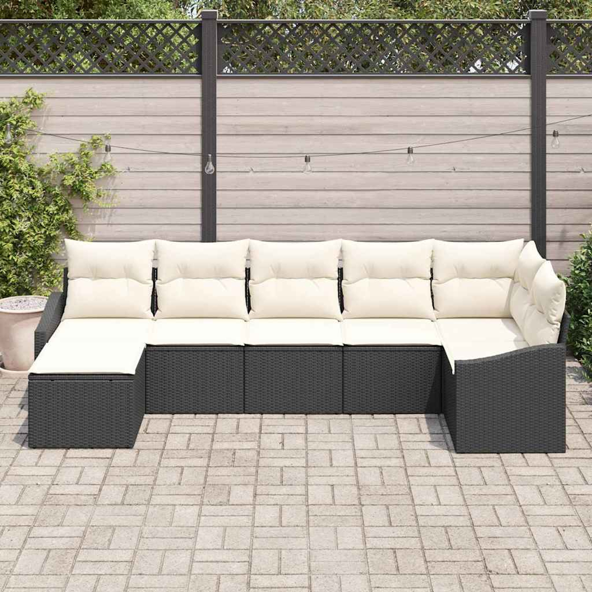 Ein dunkelgraues Ecksofa mit cremefarbenen Kissen steht auf einer Ziegelterrasse. Im Hintergrund befinden sich ein Holzzaun und Lichterketten.