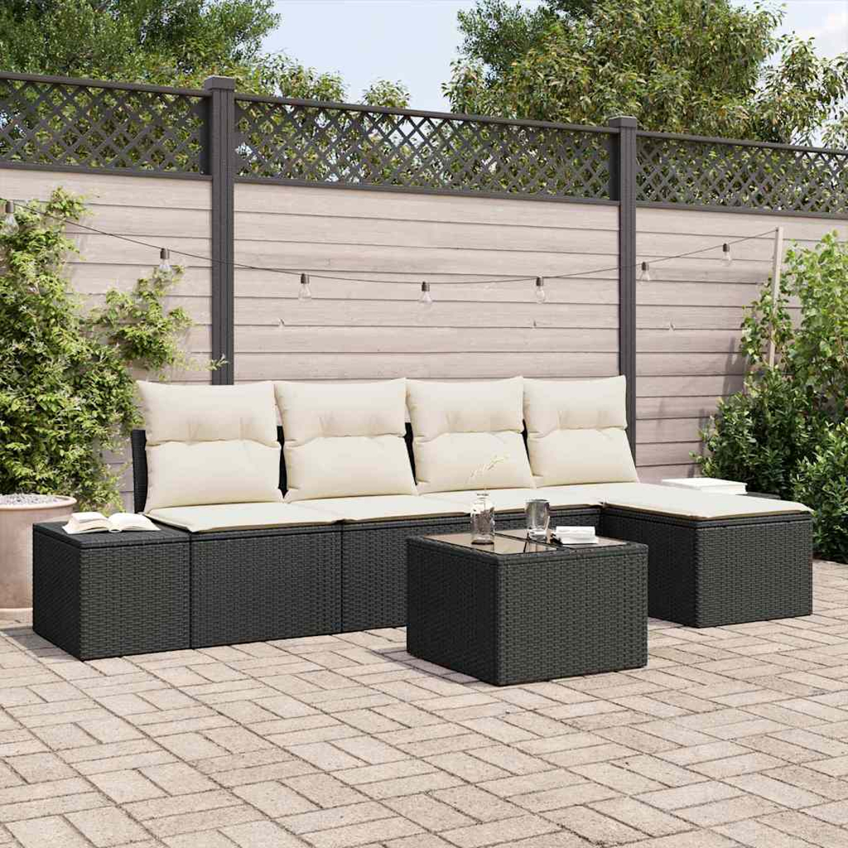 Terrassenset mit dunklen Korbmöbeln, cremefarbenen Kissen und einem Tisch mit Glasplatte. Vor einem Holzzaun.
