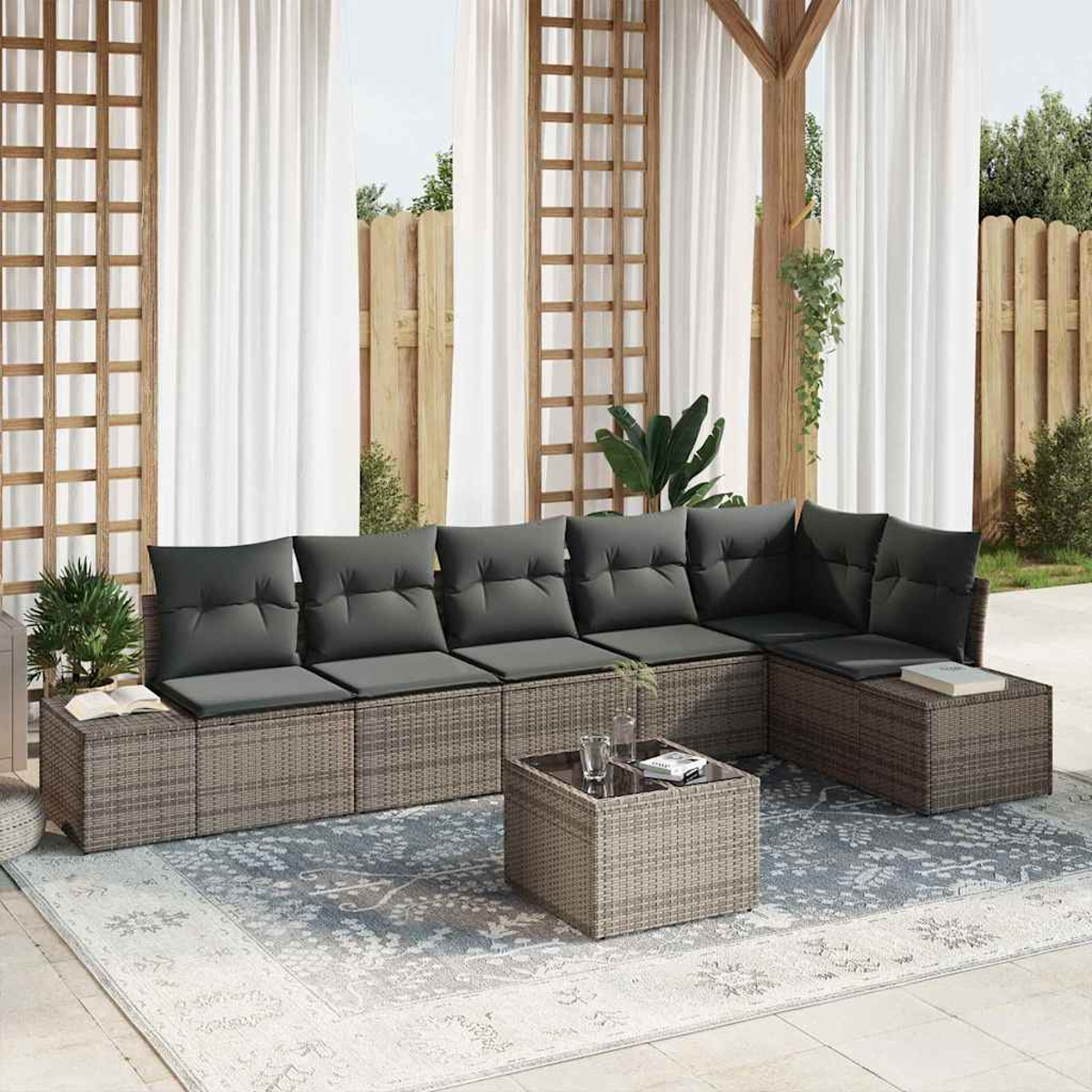 Ein Sitzbereich im Freien mit grauem Ecksofa, quadratischem Couchtisch und gemustertem Teppich. Weiße Vorhänge und Holzakzente sind sichtbar.