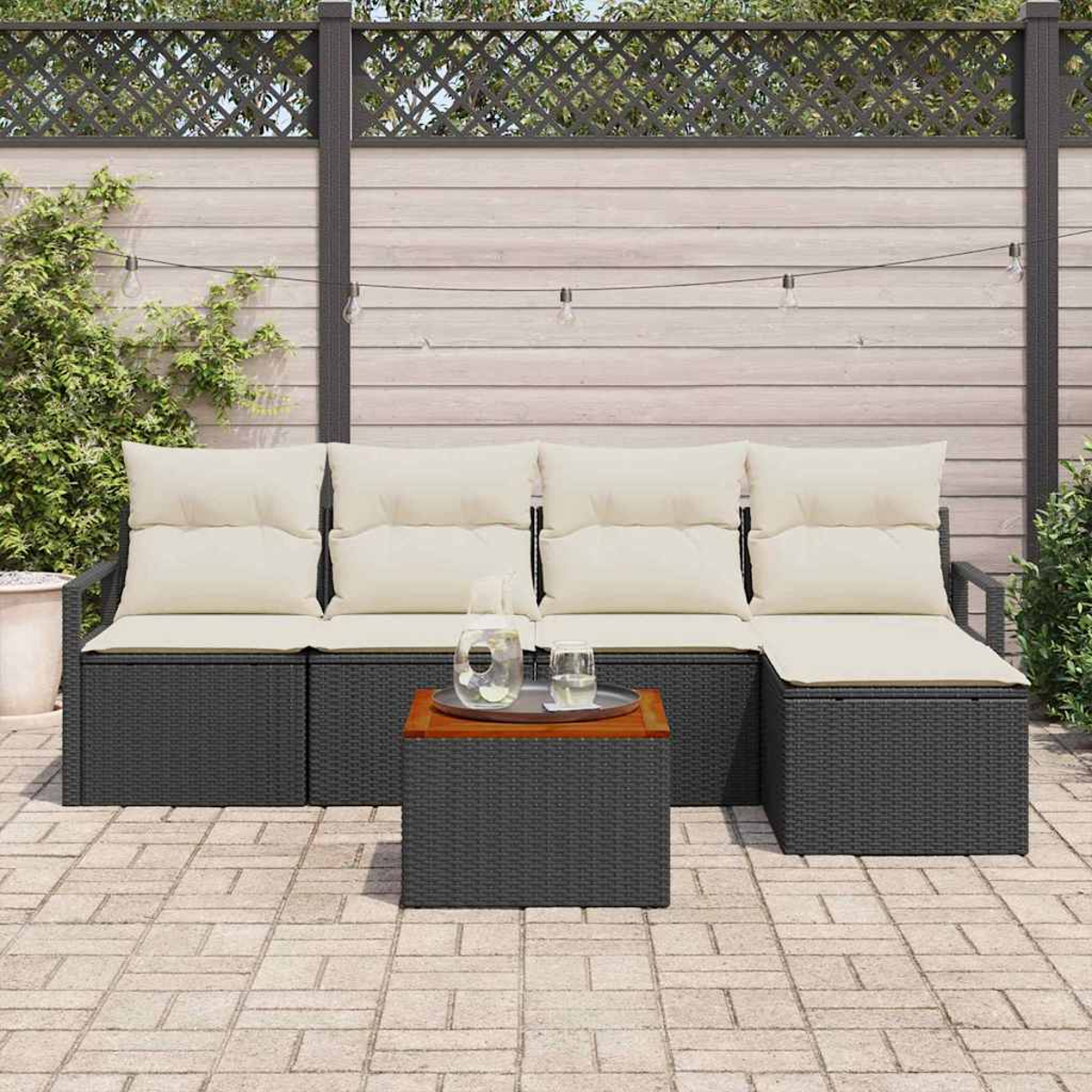 Juego de patio al aire libre con un sofá de mimbre gris oscuro, cojines crema y una mesa pequeña. Una valla de madera y plantas en el fondo.