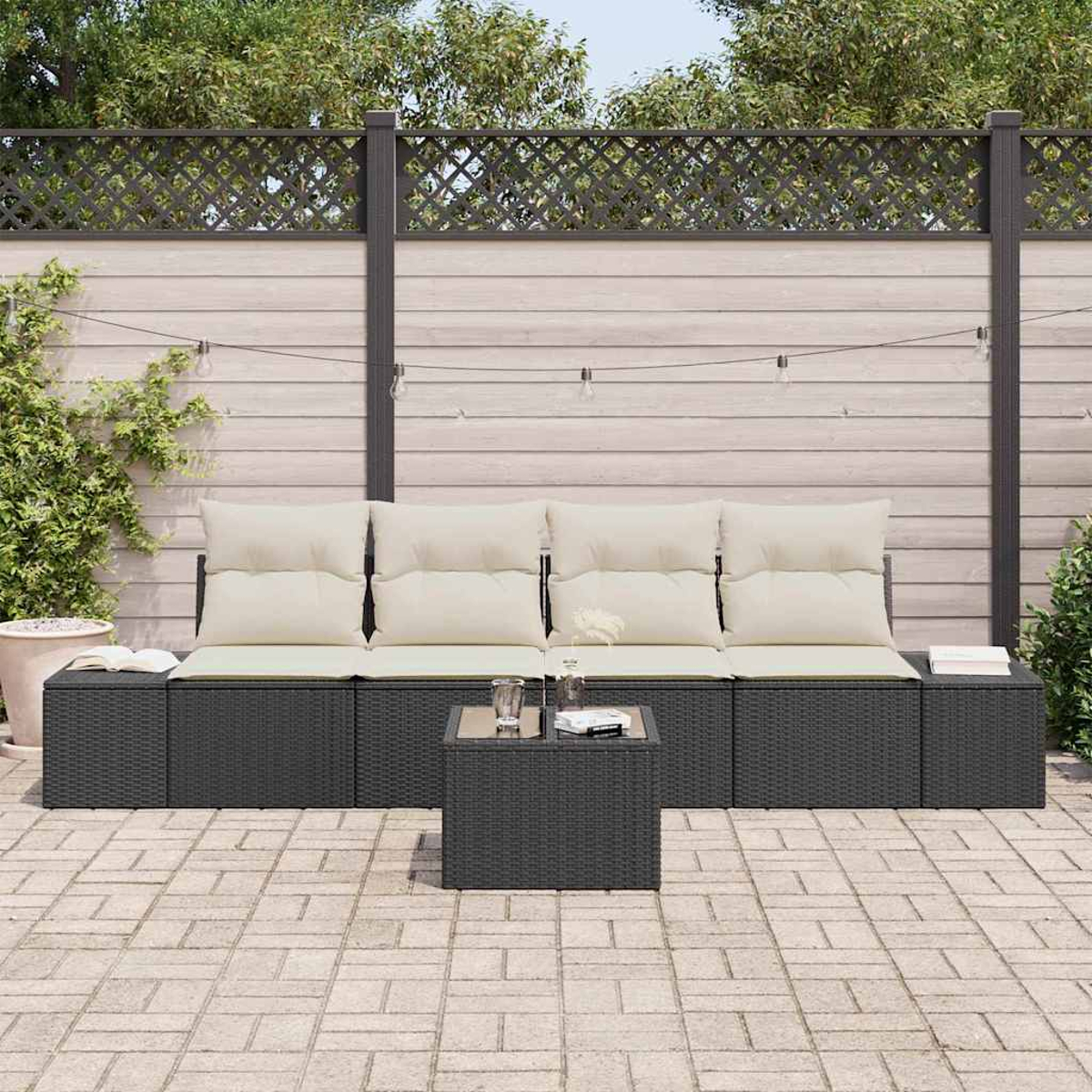 Terrassenmöbel-Set im Freien mit schwarzem Korbmöbel, cremefarbenen Kissen und einem kleinen Tisch. Ziegelstein-Terrasse mit Holzzaun und Pflanzen.