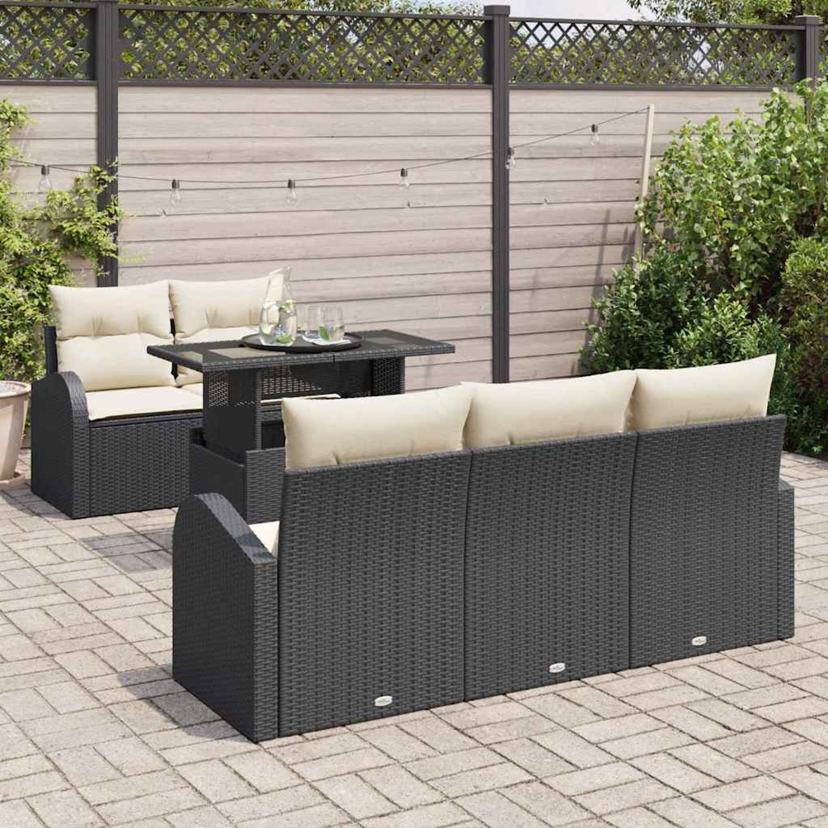 Juego de patio al aire libre con muebles de mimbre gris oscuro, cojines beige y una mesa con tapa de cristal. Contra una valla de madera.