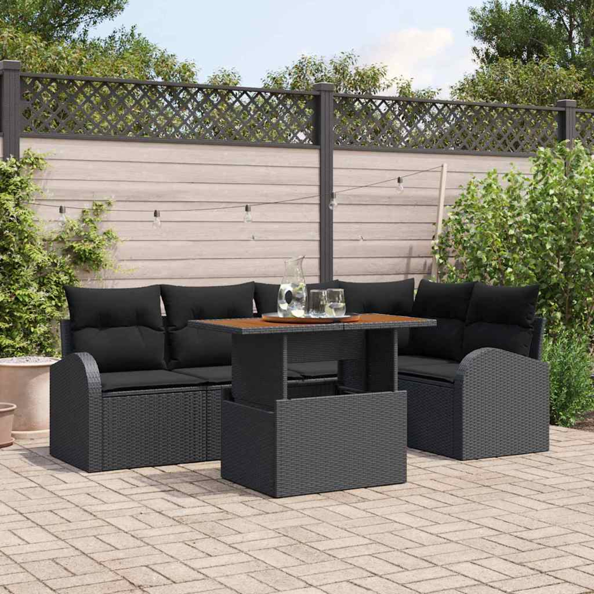 Garten-Terrassenset mit schwarzen Kissen und Holztisch. Zaun und Grün im Hintergrund. Ziegel auf dem Boden.