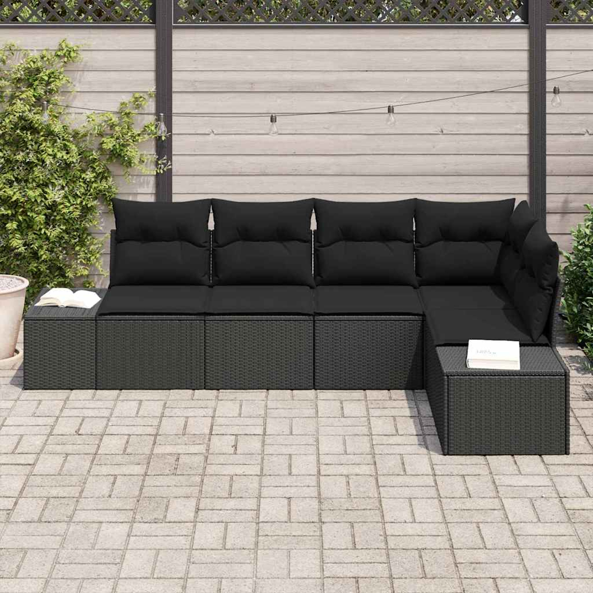 Schwarzes Korbsessel-Sofa mit Kissen auf einer Terrasse. Ein Buch liegt auf einem Beistelltisch. Holzzaun im Hintergrund.