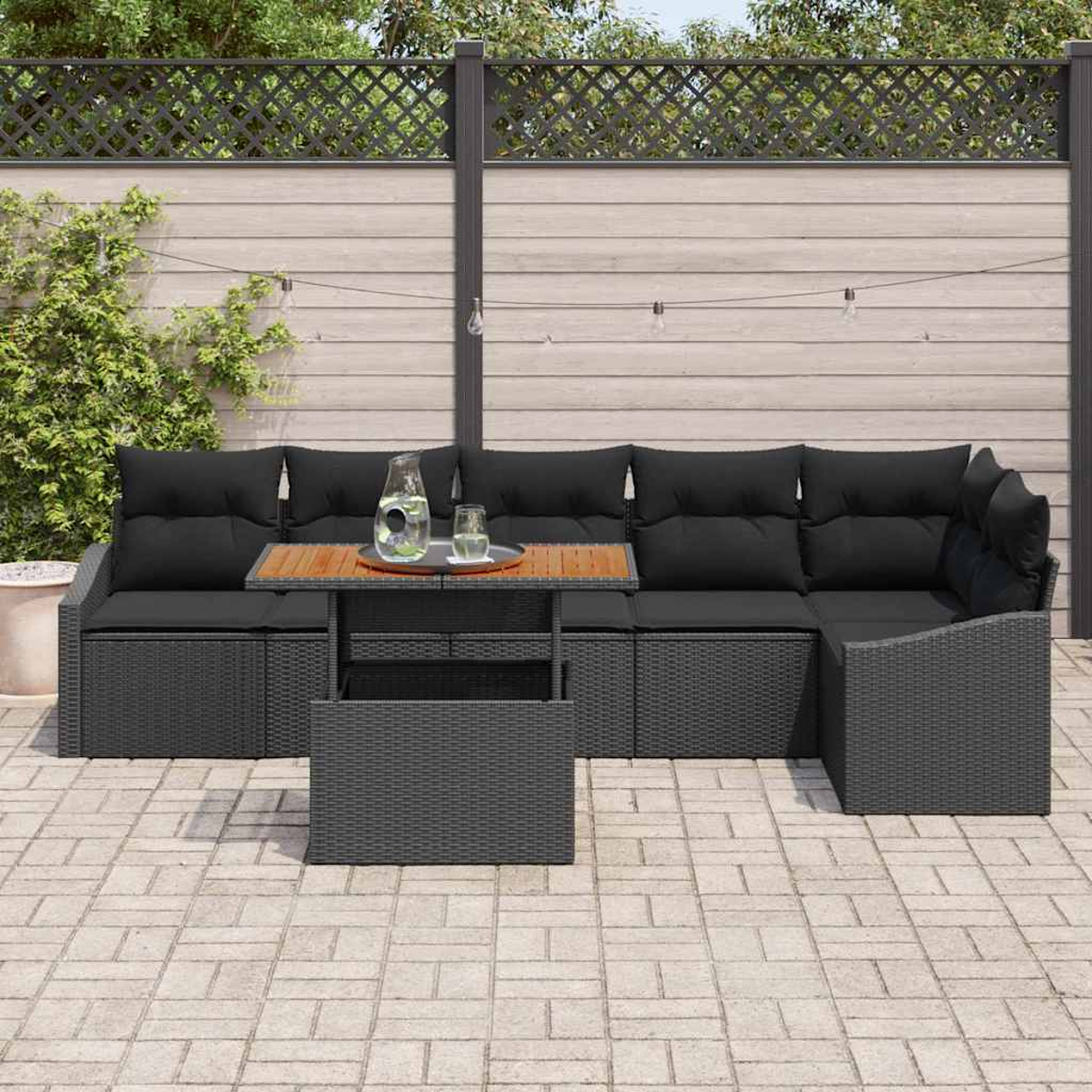 Outdoor-Ecksofa mit schwarzen Kissen. Vorne steht ein Holztisch mit Wasserkrug und Gläsern. Der Zaun befindet sich hinter dem Sofa.