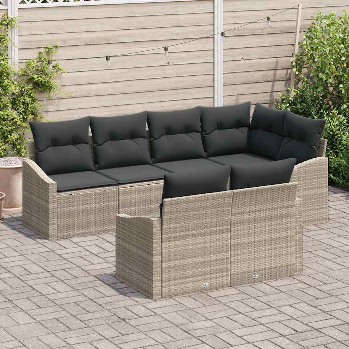 Outdoor-Sofa aus Korbgeflecht mit grauen Kissen, auf einer Ziegelterrasse, mit Holzzaun und Grün im Hintergrund.