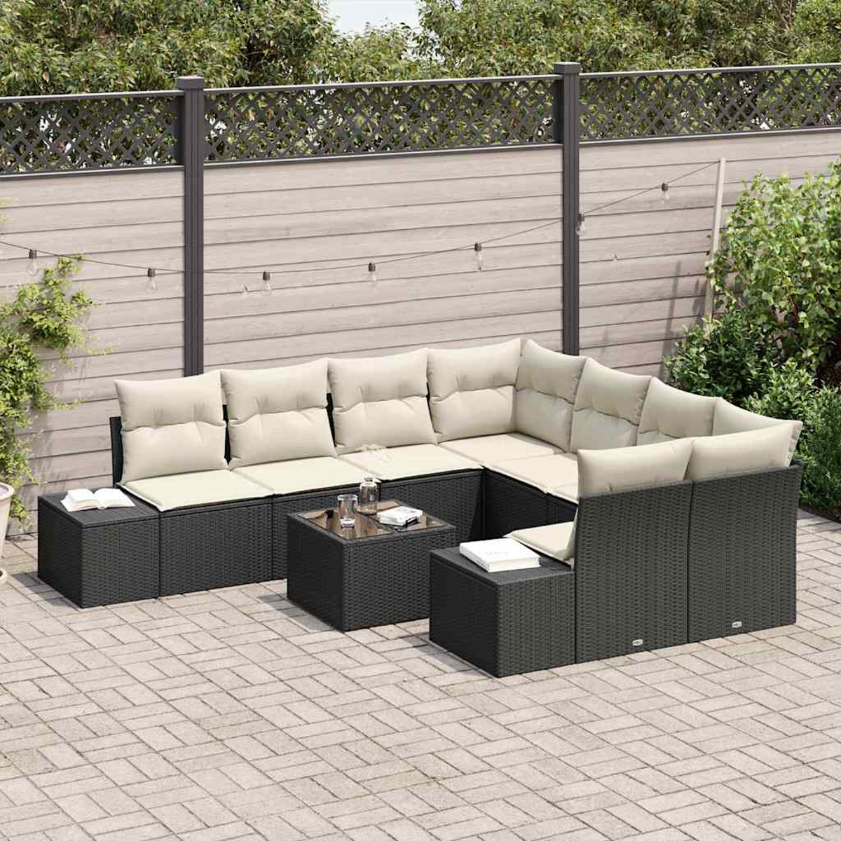 Outdoor-Ecksofa mit beigen Kissen auf einer Backstein-Terrasse. Ein kleiner Tisch trägt Gläser, Bücher. Holzzaun mit Pflanzen im Hintergrund.