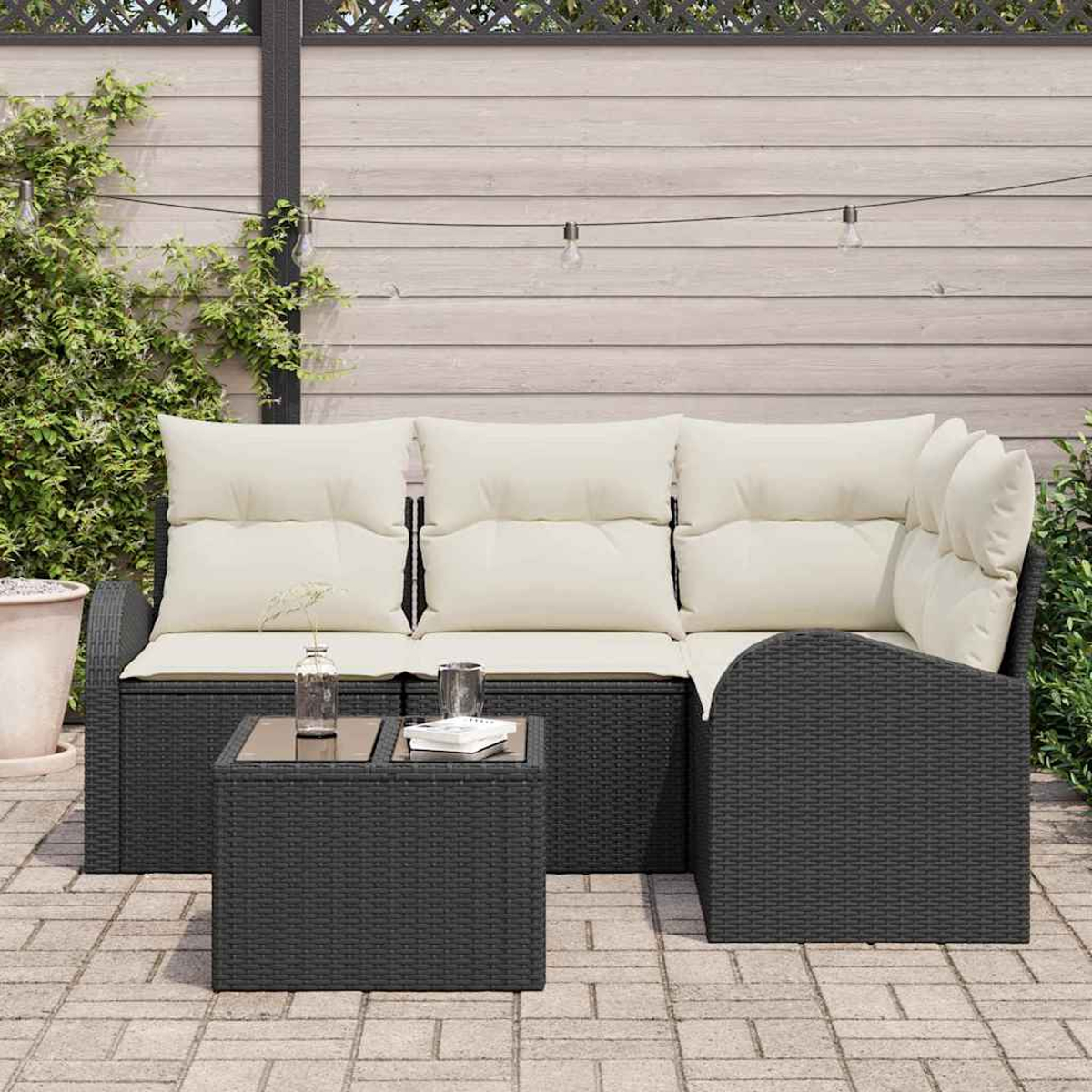 Juego de patio exterior con muebles de mimbre negro, cojines crema y una mesa pequeña. Fondo con valla de madera y luces de cadena.