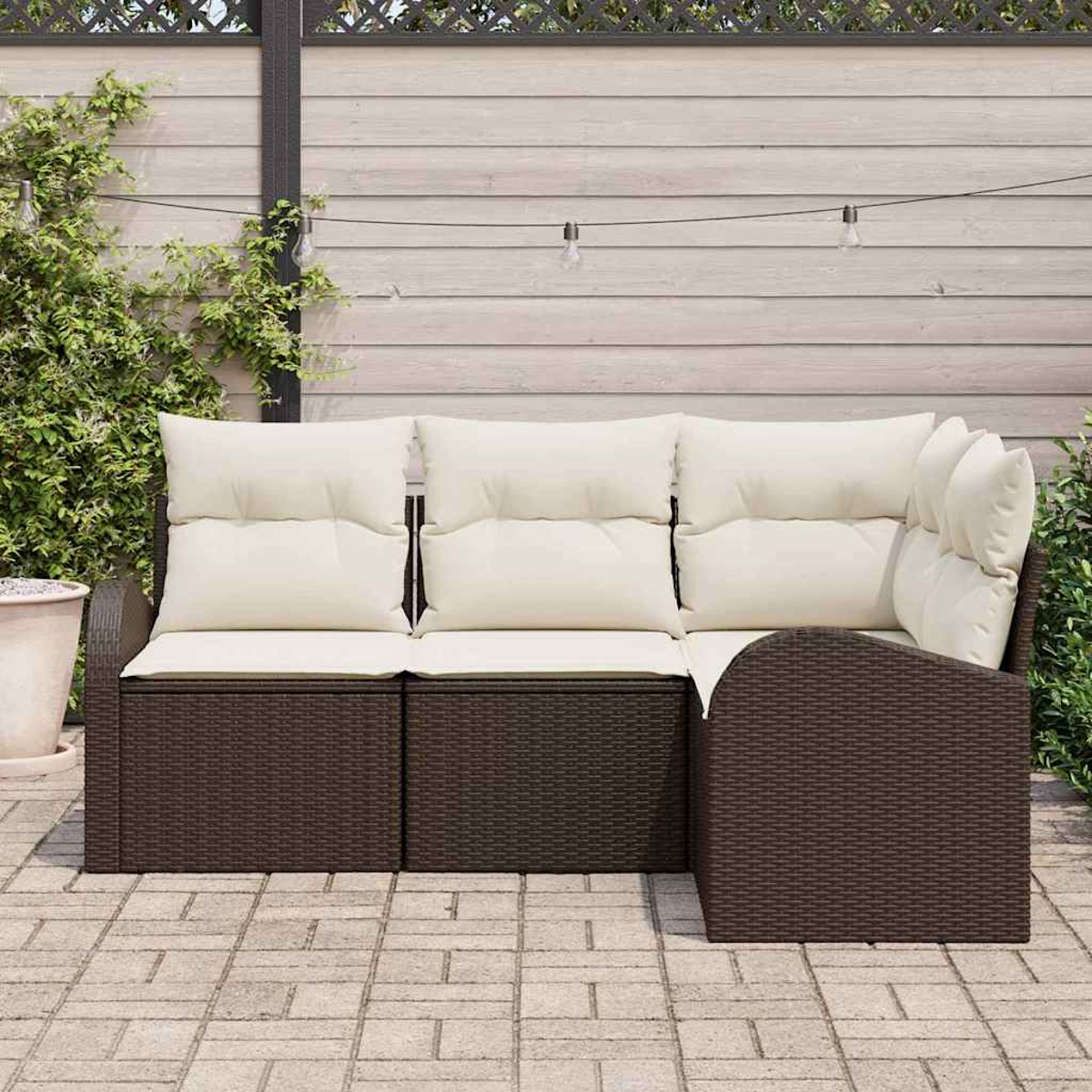 Braunes Outdoor-Ecksofa aus Korbgeflecht mit cremefarbenen Kissen, auf einer Backstein-Terrasse vor einem Holzzaun mit Pflanzen.