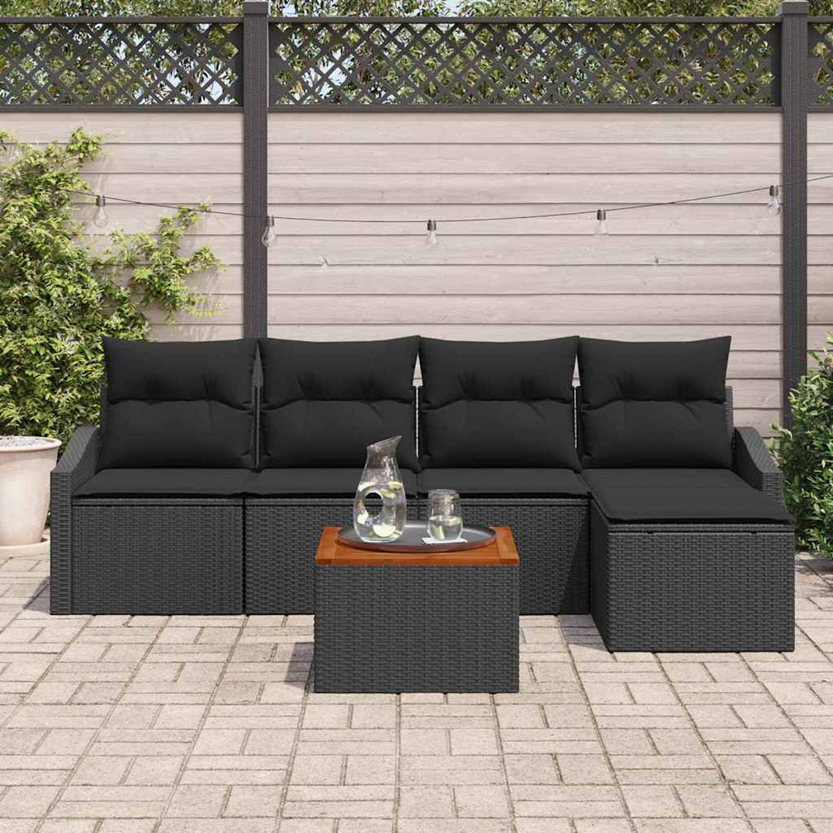 Outdoor-Ecksofa mit schwarzen Kissen. Ein Couchtisch mit Holzplatte, Glaskrug und Gläsern. Im Hintergrund ein Holzzaun.